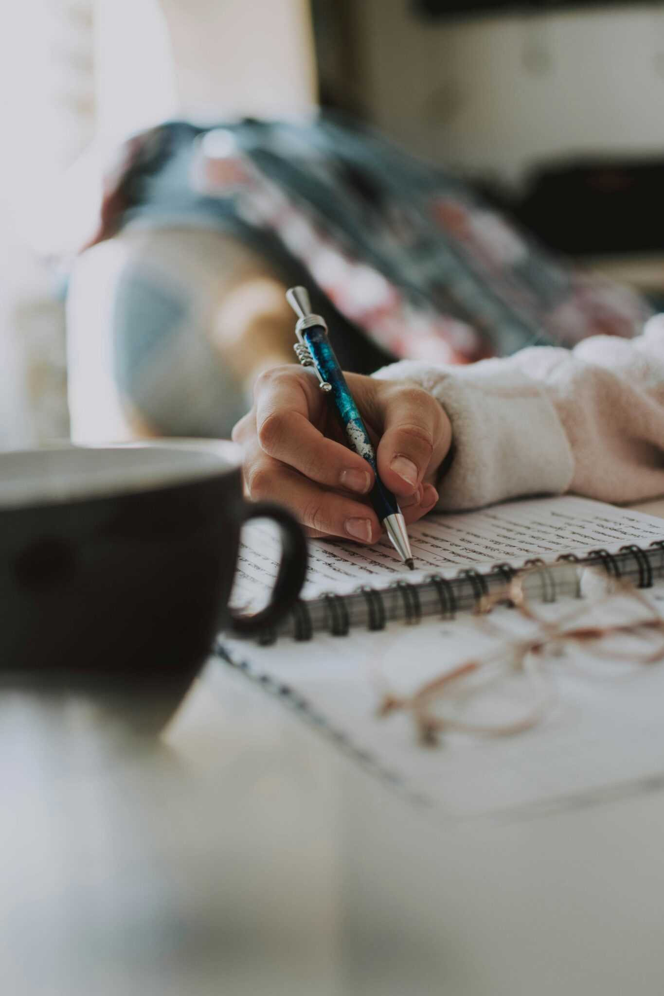 Close-up of a hand writing in a spiral notebook beside a coffee mug, reflecting on your part in repair.
